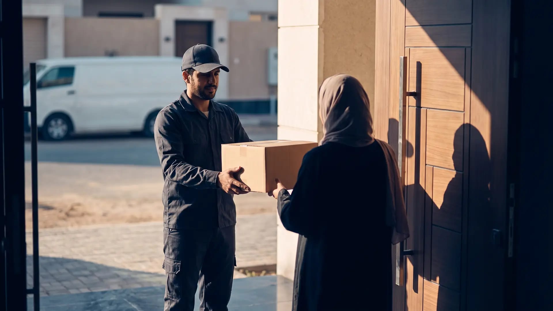 A courier in a plain uniform handing an unmarked parcel to a customer at a Riyadh doorstep in late-afternoon light