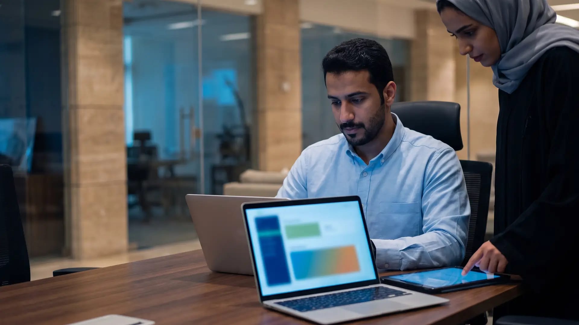 A sales manager and a colleague reviewing pipeline dashboards on a laptop and tablet in a Riyadh office