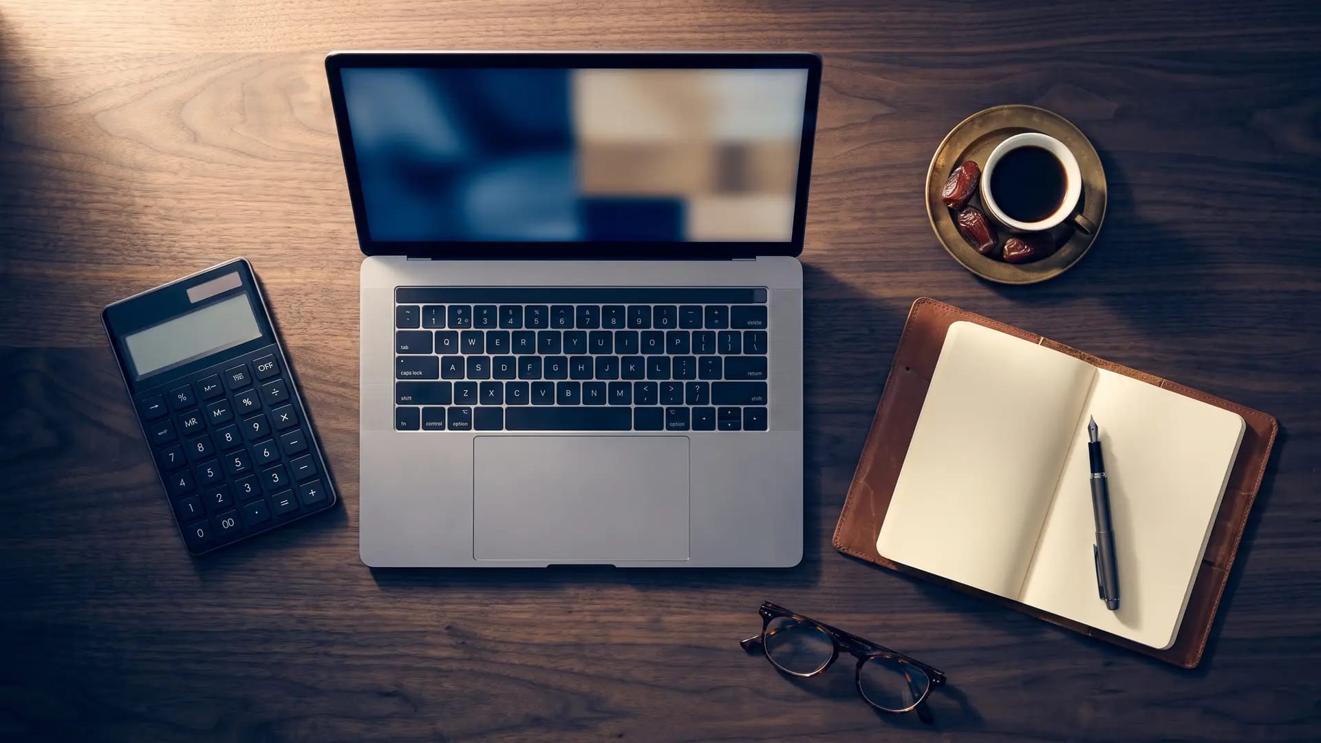 A desk laid out with a laptop, calculator, notebook and Arabic coffee for a software cost comparison