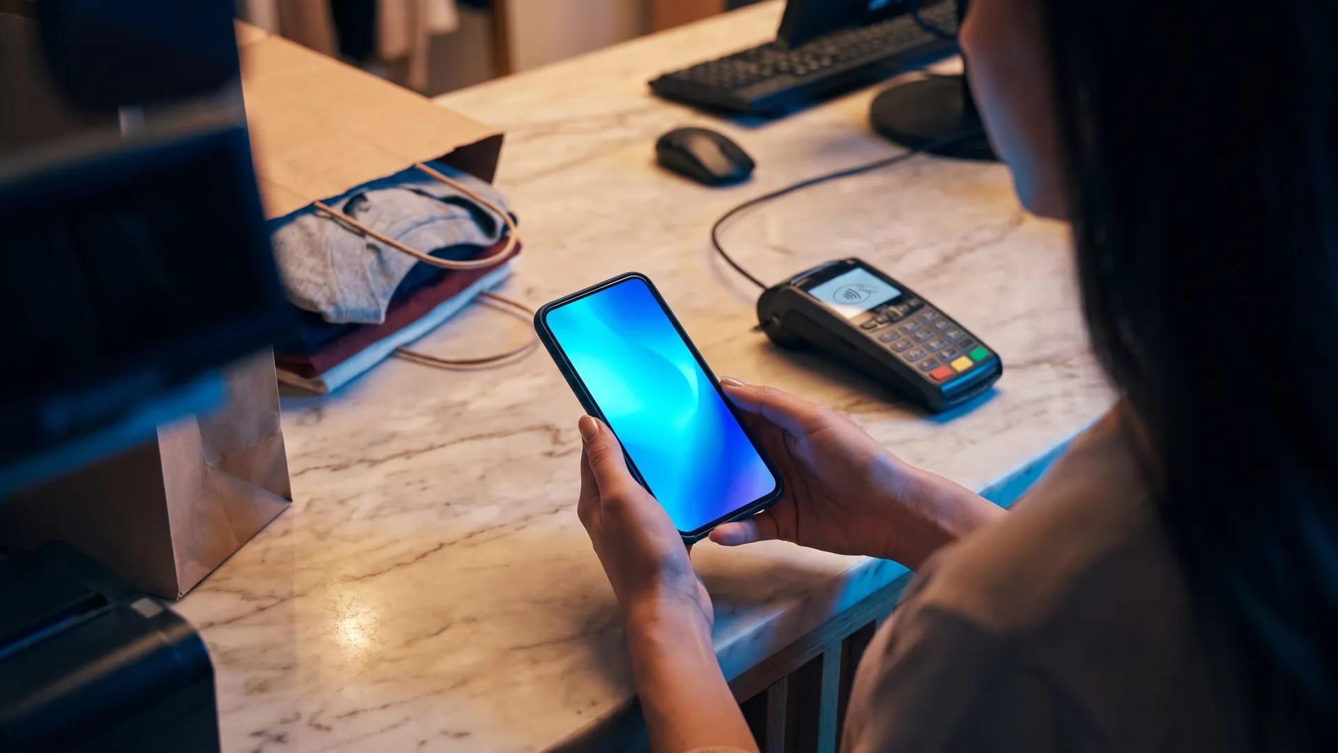 A shopper holding a smartphone at a retail checkout counter beside a contactless card terminal and a paper bag